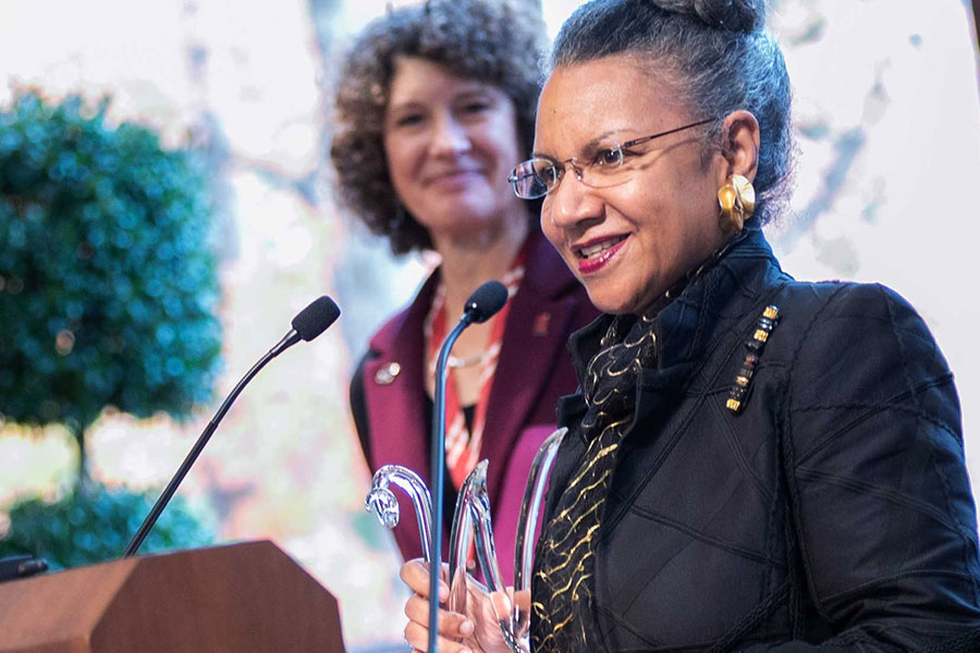 A woman stands at a podium holding a glass award and smiling as she speaks into a microphone, while another woman stands behind her listening; the setting appears to be a formal Women’s Philanthropy event.