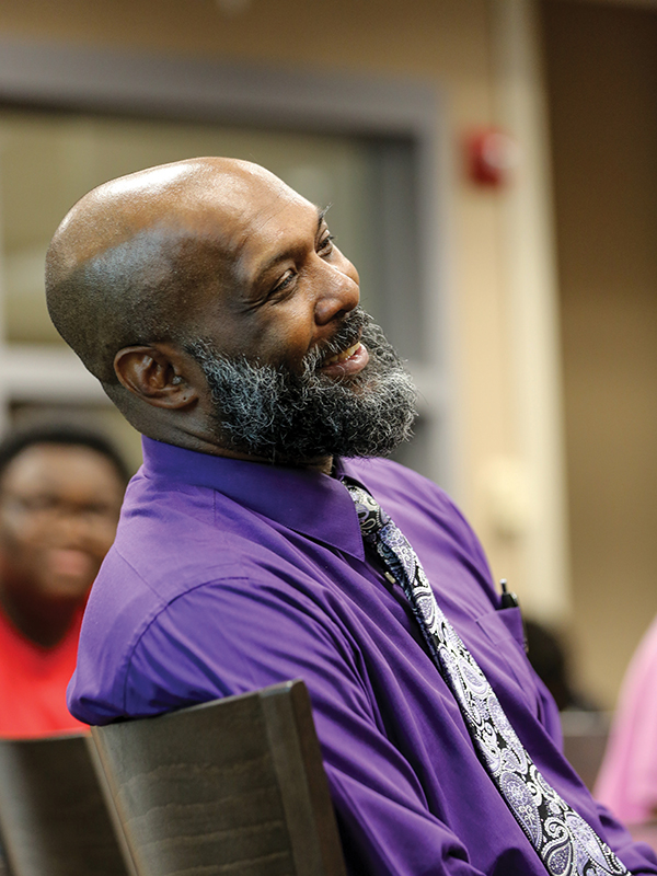 Sherman Woodard, wearing a purple button-up shirt and tie, sits back in his chair and smiles. In soft focus behind him is a high school student smiling. 