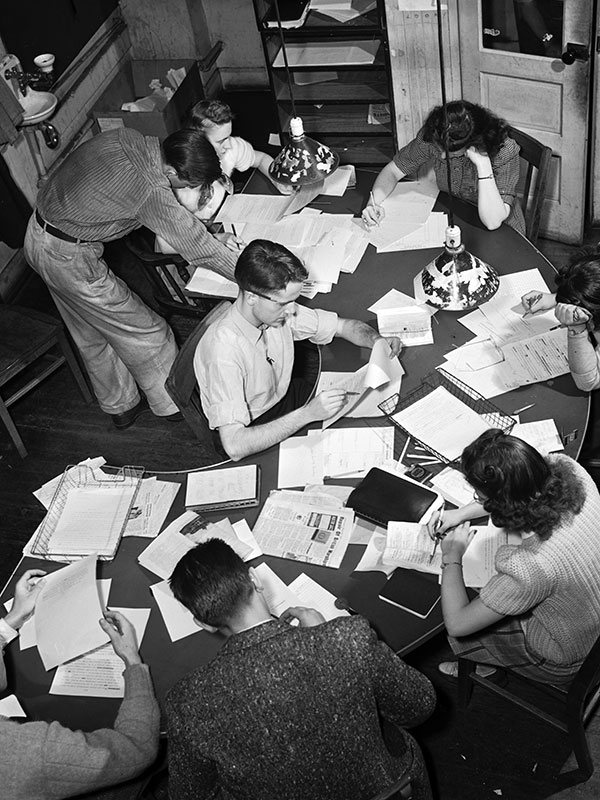 A black-and-white photo shows students huddled over a crescent shaped table, engrossed in their work, papers strewn about everywhere.