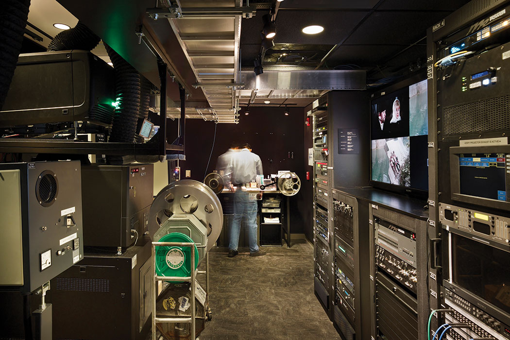 A person, blurred in movement, works in the projection booth of the IU Cinema. Brightly lit, the booth features wall-to-wall, floor-to-ceiling technology, including two film projectors to accommodate both 35mm and 16mm film reels, as well as two digital projectors and a touchscreen computer to orchestrate each cinematic event.