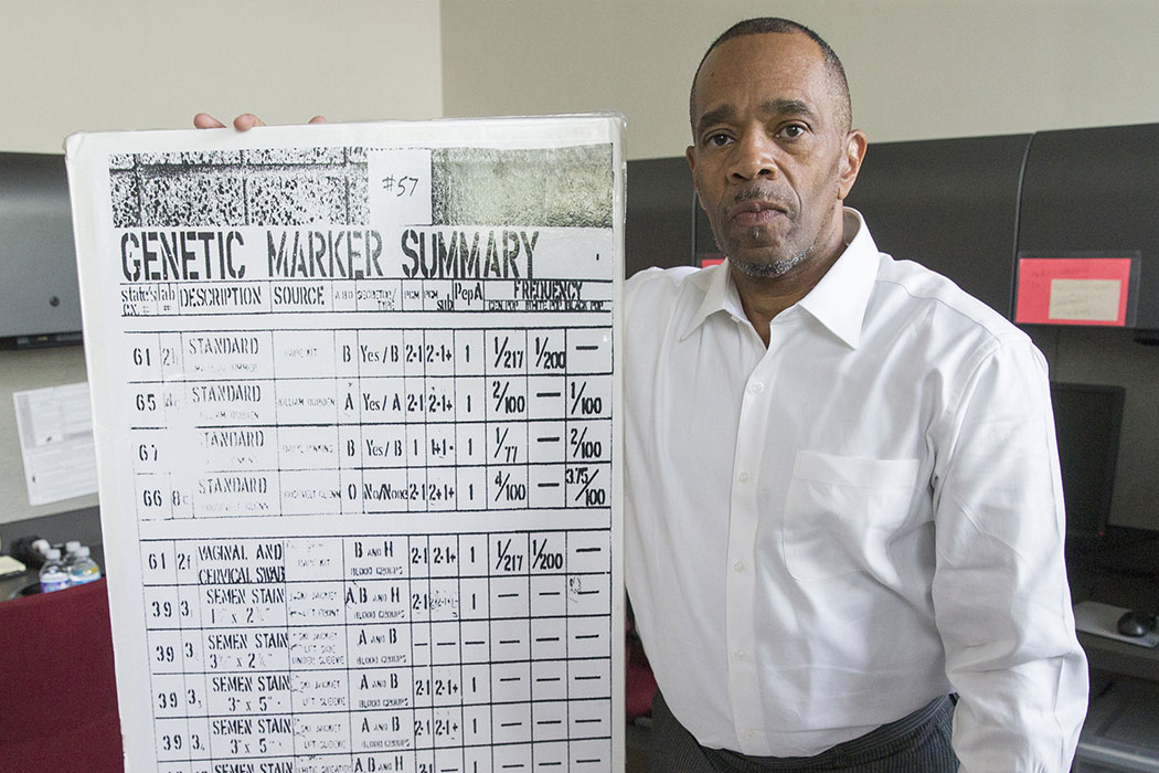 Pinkins, in a crisp, white button-up shirt, holds a poster-size enlargement of a black-and-white report. It says "Genetic Marker Summary" and has several rows and columns marked with numbers and labels.