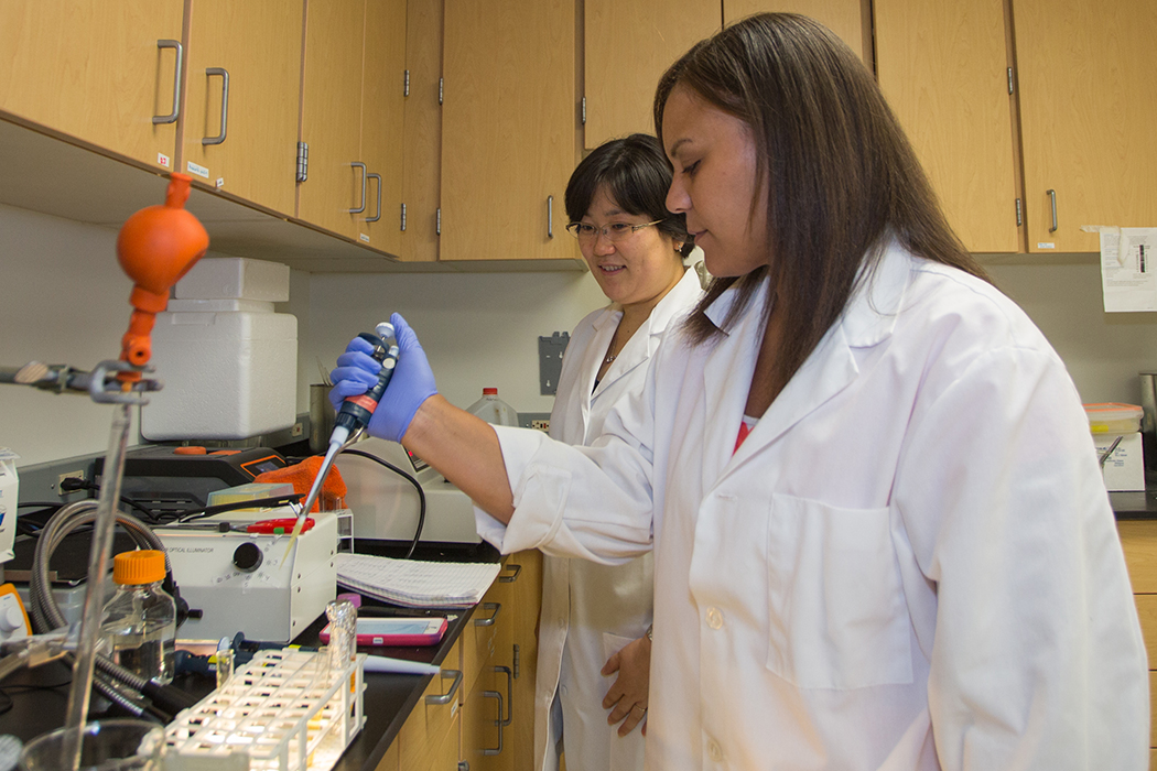 Kortany Baker, wearing a white lab coat and blue rubber gloves, uses scientific equipment to test bacterial solutions. Hisako Masuda, also in a white lab coat, oversees.