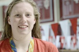 Lilly King, wearing an Olympic gold medal around her neck, smiles. She stands in front of a blurred background of banners touting the many accomplishments of IU Swimming and Diving over the years.