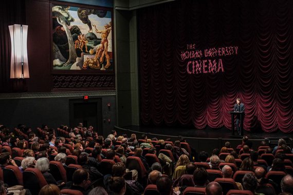 A man in a dark suit stands before a full, seated audience. He stands at a podium on an elevated stage, lit by a spotlight in a dimly lit theatre. Behind him, the words “The Indiana University Cinema” is projected on the richly hued, velvet curtains that are gathered in luxurious folds.