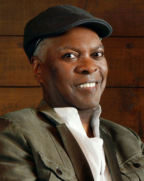 Booker T. Jones, wearing a white button-up shirt, green jacket, and black newsboy-style cap, smiles at the camera.