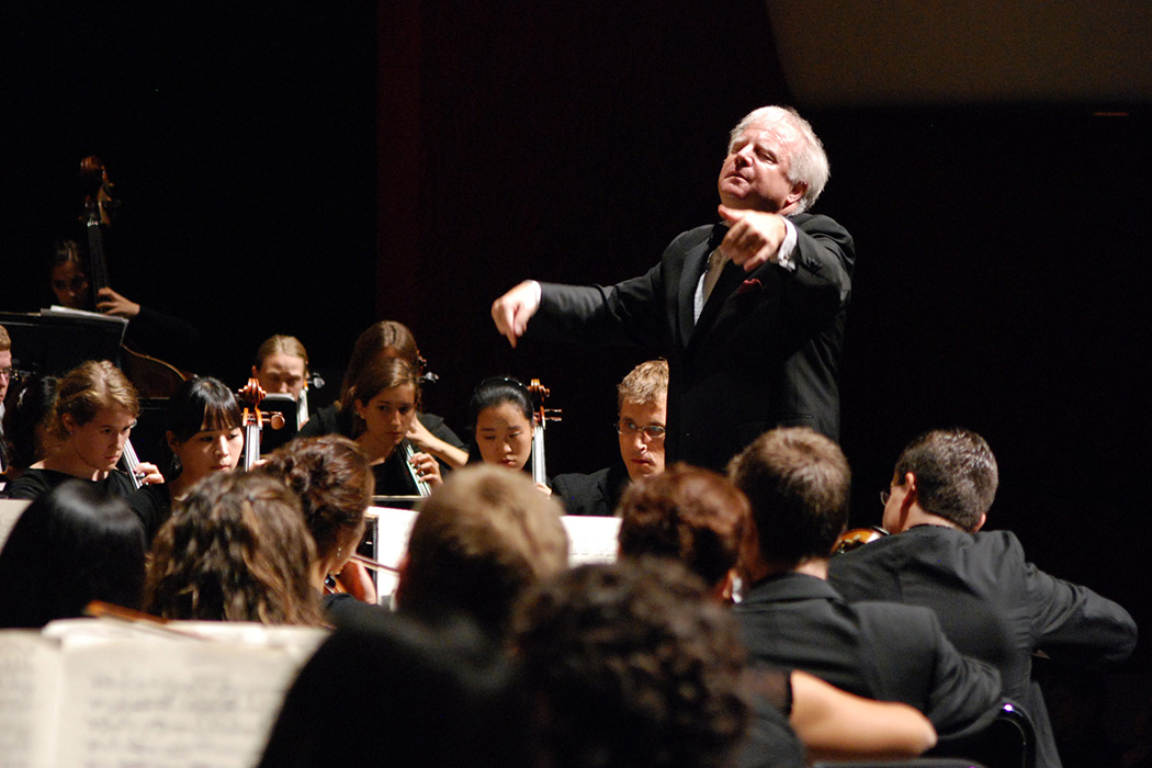 With eyes closed and arms outstretched, Leonard Slatkin stands on a raised platform conducting an orchestra.