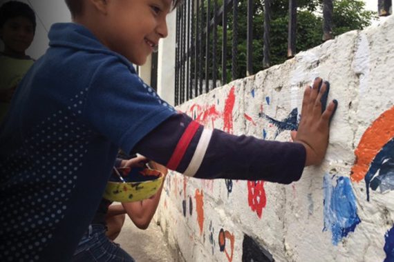 A smiling boy presses his paint-covered palm against a stone wall to mark his hand print.