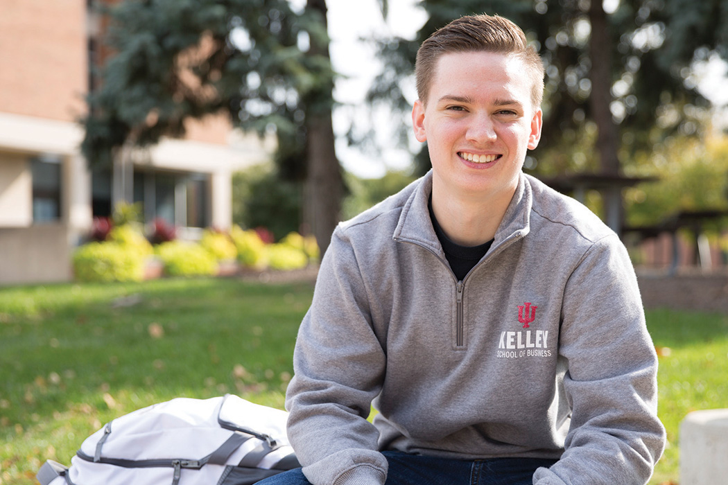 Alex Bajzatt sits outside on a campus bench, smiling at the camera while wearing a Kelley School of Business sweatshirt.
