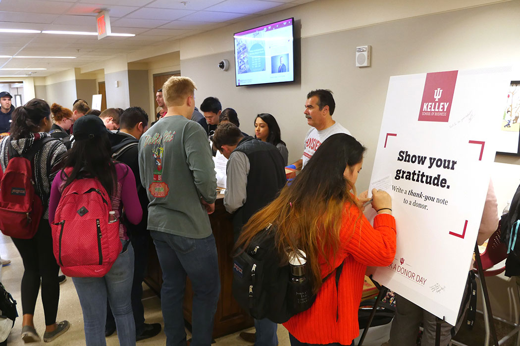 Students crowd around a table to sign thank you cards to Kelley School of Business donors. In the foreground, a woman in a bright red sweater is writing a note on a large sign that reads: "Show your gratitude. Write a thank-you note to a donor."