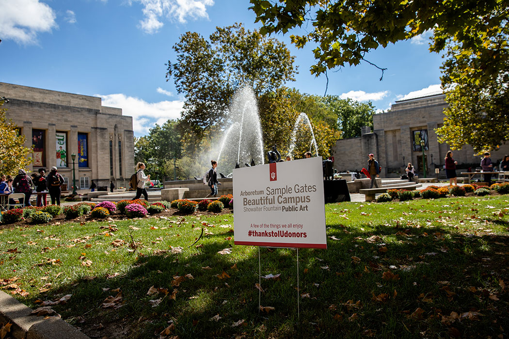 Students walk around Showalter Fountain, which sprays gloriously toward the blue sky. The limestone facades of the Lilly Library and IU Auditorium are in the distance. In the foreground, a sign placed in the ground reads: "Arboretum, Sample Gates, Beautiful Campus, Public Art, Showalter Fountain—a few of the things we all enjoy #thankstoIUdonors."