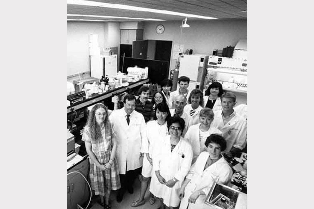 A retro black-and-white photo showing a group of people in a lab, smiling at the camera. Some of them wear white lab coats. 