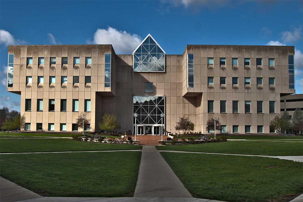 The University Library stands at the endpoint of several walkways separated by wide patches of green grass. The building is made of light stone and features many windows on its facade, including an expansive glass wall spanning several floors at its center.