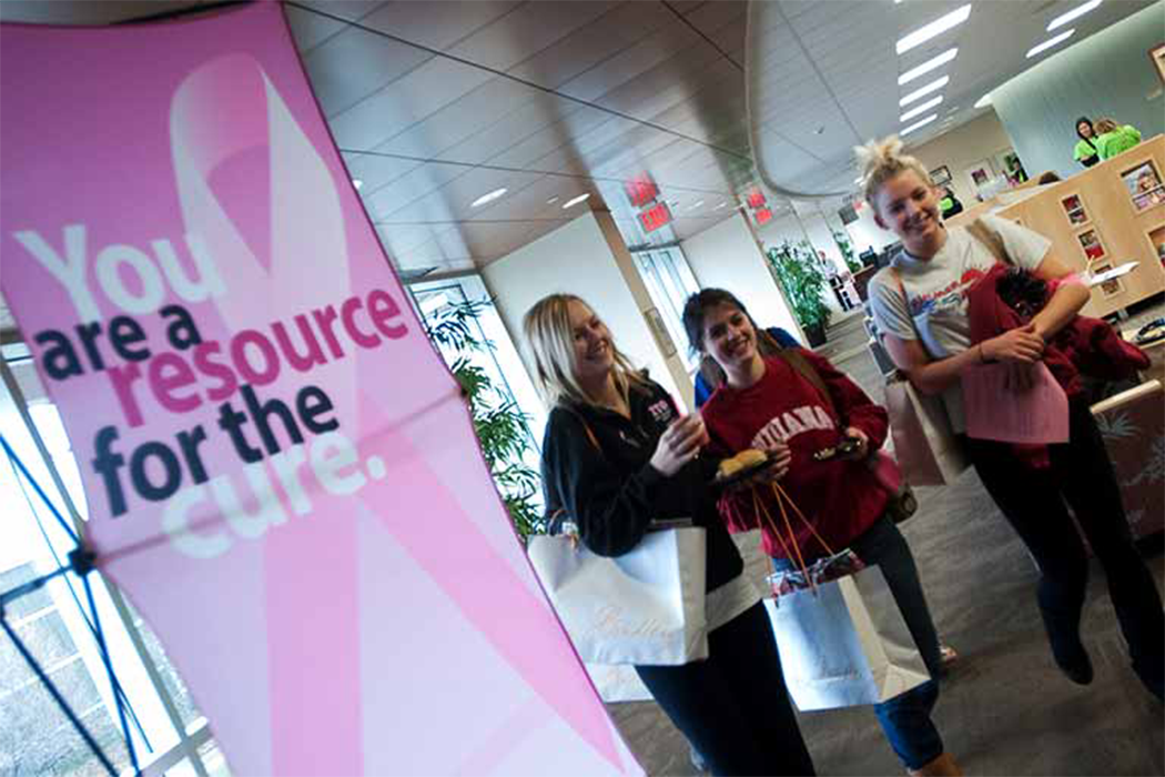 Three IUPUI students walk past a pop-up sign featuring a pink breast cancer ribbon and the headline, "You are a resource for the cure."