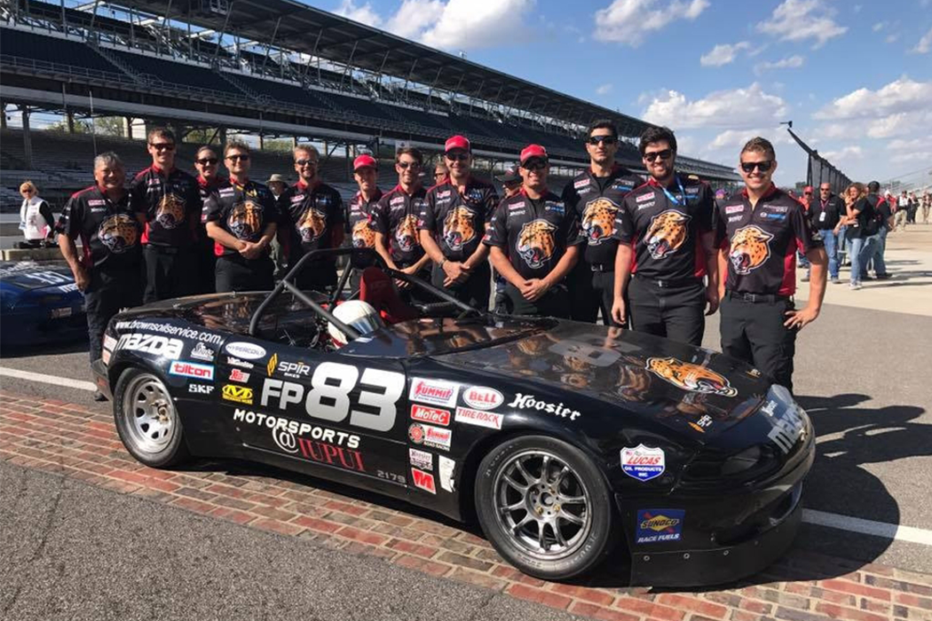 A group of people wearing matching black shirts with the IUPUI Jaguars logo stand behind a black race car at the Indianapolis Motor Speedway.
