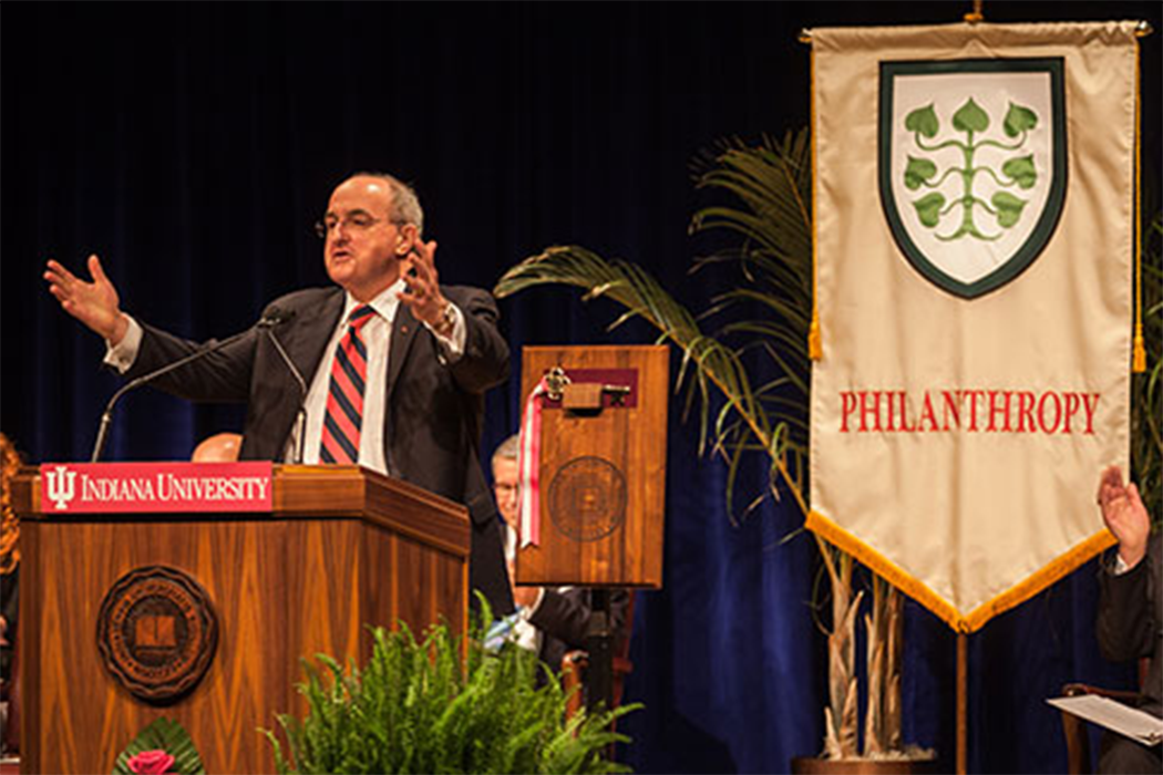 Michael McRobbie speaks from behind a podium; Behind him hangs a large banner with the word "Philanthropy" and a crest of interlocking leaves on it.
