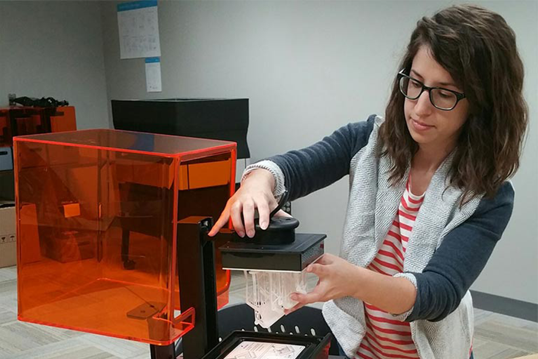 A brunette woman wearing glasses holds on to the arm of a 3D printer as it produces a palm-sized white plastic sculpture. 