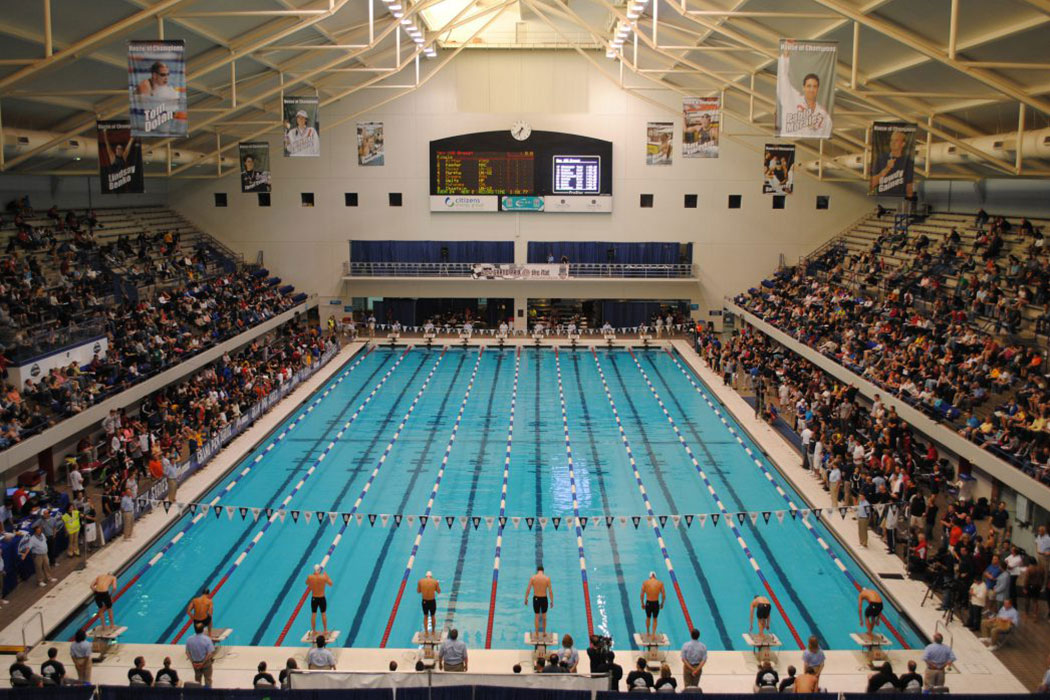 A wide shot of the IU Natatorium swimming pool shows swimmers lining up on the blocks, as the crowd in the stands on either side of the pool look on. 
