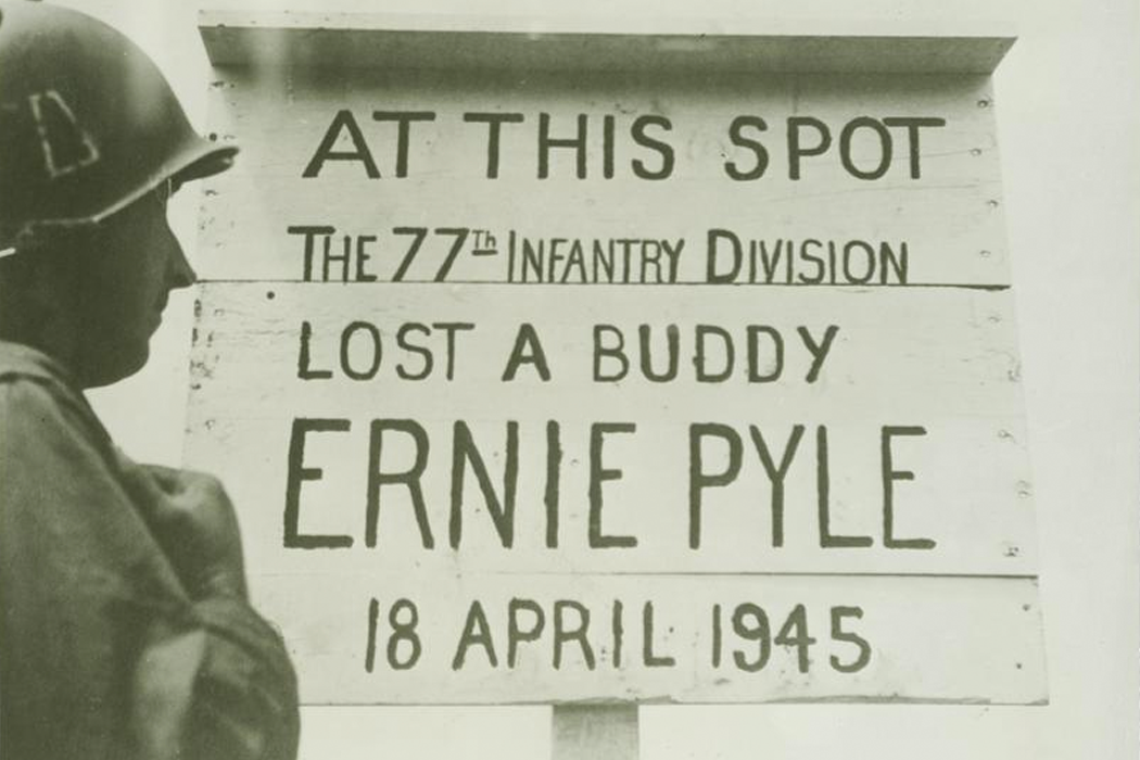 Black-and-white photo of a soldier reading a makeshift memorial for Ernie Pyle that reads, "At this spot the 77th infantry division lost a buddy: Ernie Pyle, 18 April 1945."