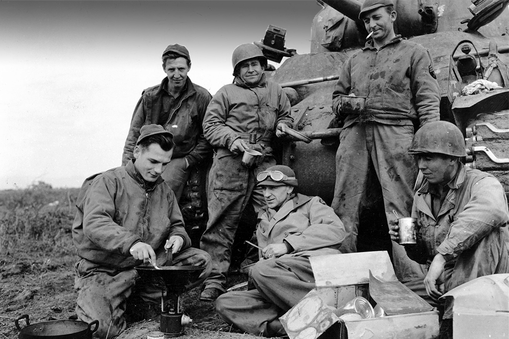 Black-and-white photo of Pyle and a group of soldiers relaxing in front of a tank while heating field rations in a skillet.
