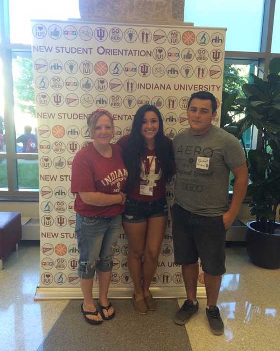 Three people stand in front of a banner with graphics all over it and the words "Indiana University New Student Orientation."