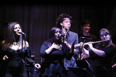 Five members of IU Soul Revue, all wearing black, perform on stage in a dimly lit venue.