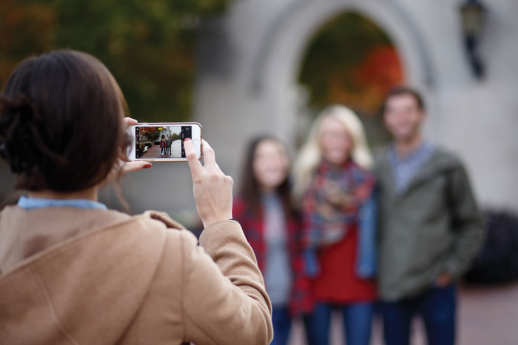 An over the shoulder shot of a woman holding a cell phone and taking a picture of two women and a man posing in front of a building.