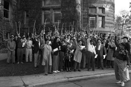 Black-and-white photo of a large group of male students in suits and hats. They are holding canes and raising them in the air as they shout.