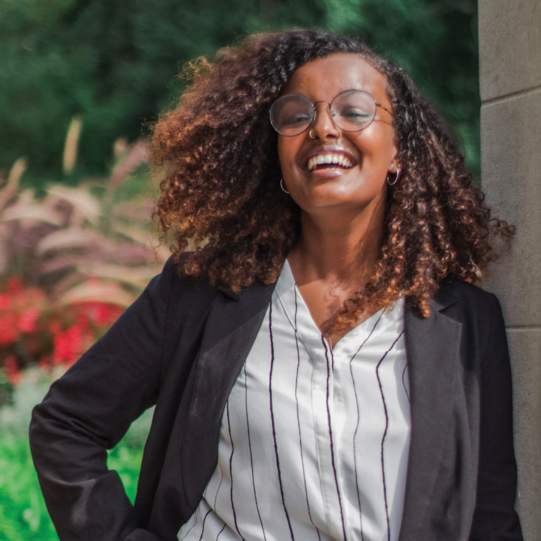 A woman with brown skin and shoulder-length, curly, brown hair lifts her chin to the side and smiles broadly. She is wearing a black blazer over a white button-up shirt with pinstripes. She has a nose ring, small hoop earrings, and round, metal-framed eyeglasses. 