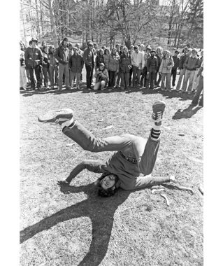 Black-and-white photo of a man with shaggy, dark hair and a thick mustache laying on the ground with his arms splayed beneath him and his legs in a V in the air. A crowd of onlookers watches from a distance.