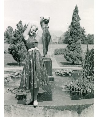 Black-and-white photo of a smiling woman with shoulder-length blonde hair. She wears flat, lace-up shoes and a sleeveless, below-the-knee dress that’s belted. The woman has her arms crossed behind her head, mimicking the pose of the nude female statue in the garden behind her.