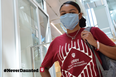 A student with medium-brown skin wearing an IU shirt and light-blue face mask holds onto the strap of their backpack and looks at the camera.