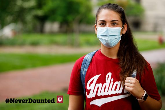 A student with brown hair in a ponytail, and wearing a red Indiana T-shirt and a light-blue face mask, walks through the Bloomington campus.