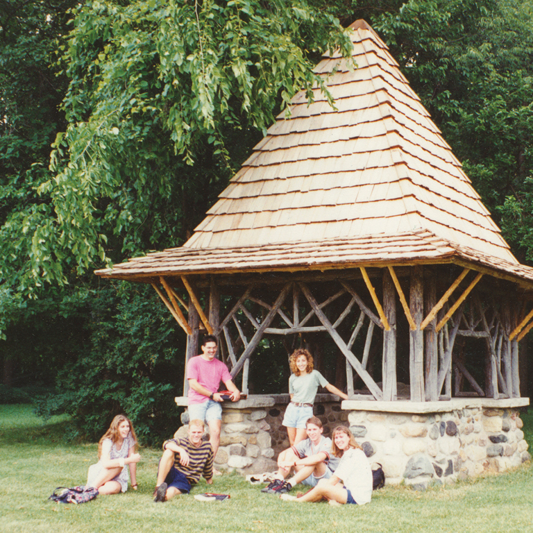 A group of six students pose in front of the Well House (a structure with a stone base, open frame of natural wood, and peaked roof of shingles) which sits along a thicket of trees.