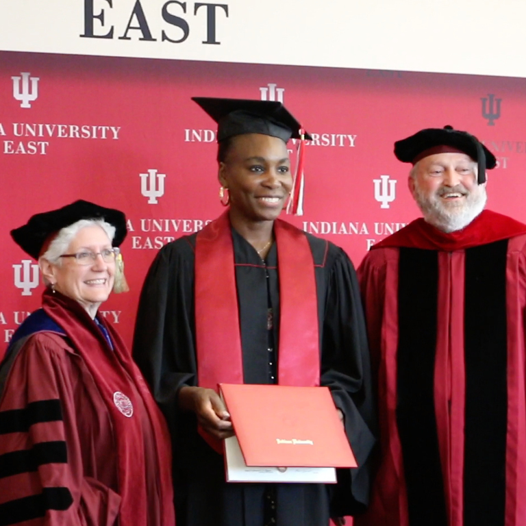 Venus Williams, a tall Black woman wearing a graduation cap with tassel, black graduation robe with red stoll, and holding a red diploma holder. On either side of her are an older white woman and older white man wearing academic regalia.