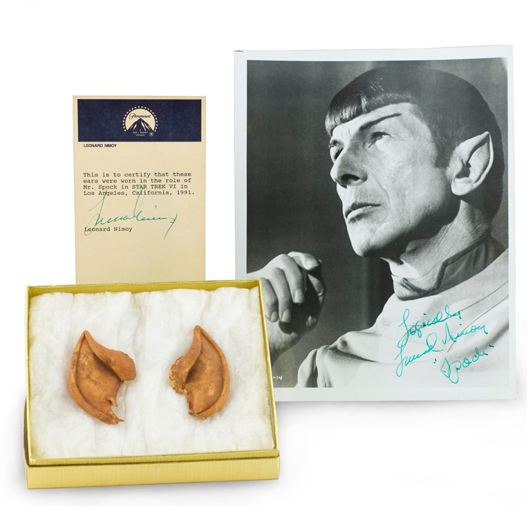 Two tan, pointy-tipped ears sit in a lined, golden box next to a signed photograph depicting Leonard Nimoy as the character Spock, and a certificate of authenticity from Paramount.