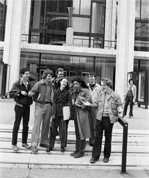 Black-and-white photo of seven college students in early 1980s garb, standing on steps in front of a building with a glass facade.