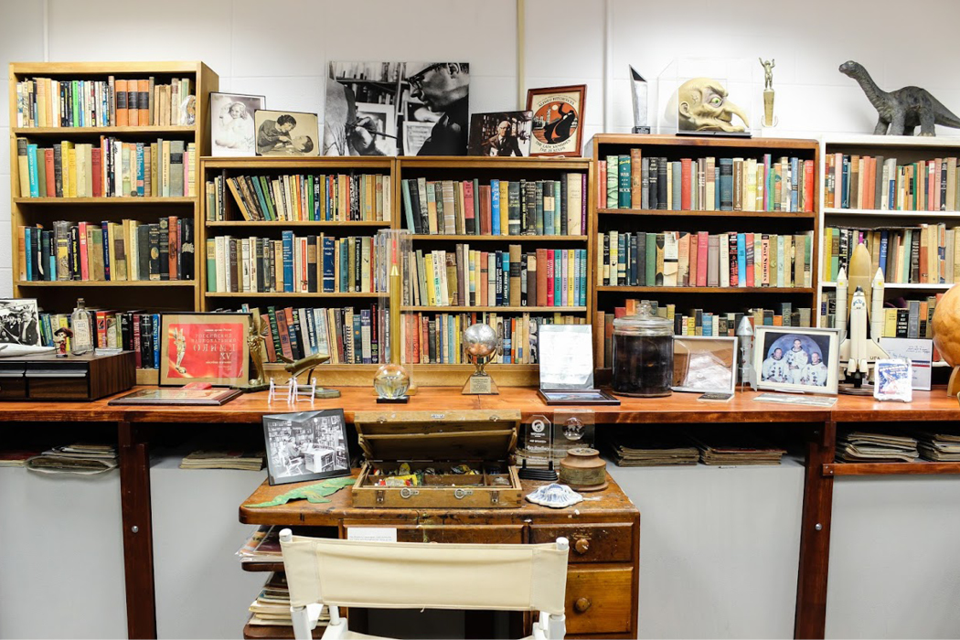 Wooden shelves packed with books and other ephemera, such as a miniature dinosaur, monster mask, framed pictures, awards, and more.