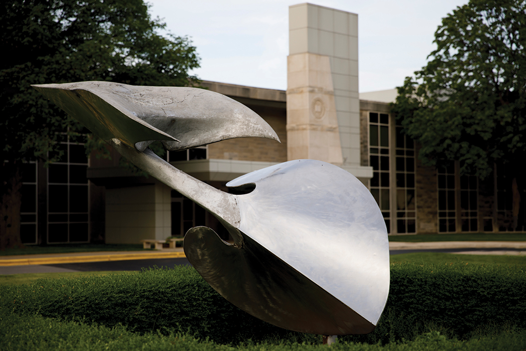 A silver sculpture that looks somewhat like a spade atop the grass, tilted 45 degrees, with a pointy beak affixed to the top.