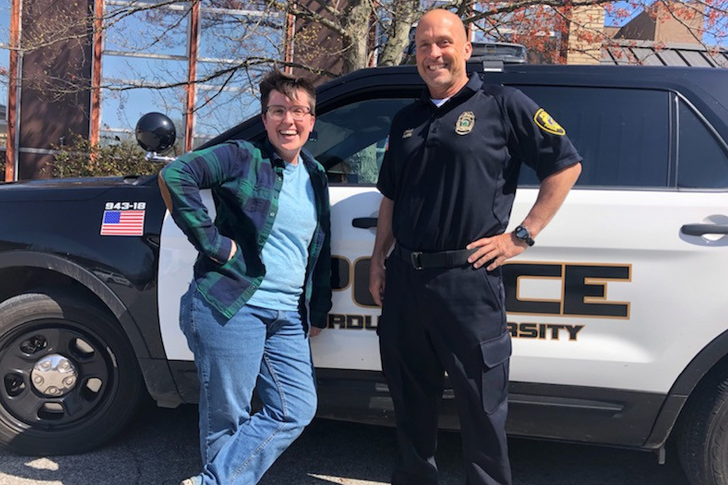 Two people stand in front of a police vehicle. On the left is a person with short brown hair, wearing glasses; an unbuttoned, long-sleeve, blue-and-green plaid shirt over a light-blue T-shirt; blue jeans; and sneakers. They have their right hand on their hip and they're leaning against the vehicle. The person on the right is bald and stands with his left hand on hip. He wears a black, short-sleeve, collard shirt with a police logo on it; black cargo pants; and black shoes.