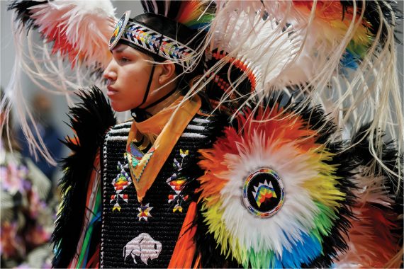 A person in powwow regalia that includes weaving, beadwork, and feathers of red, green, orange, yellow, and blue.