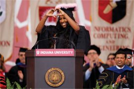 Standing at an Indiana University podium stands a woman with brown skin and long. dark hair and wearing a black graduation gown and mortar board cap with pink feathery decor on top. She is smiling and making the shape of a heart with her hands.