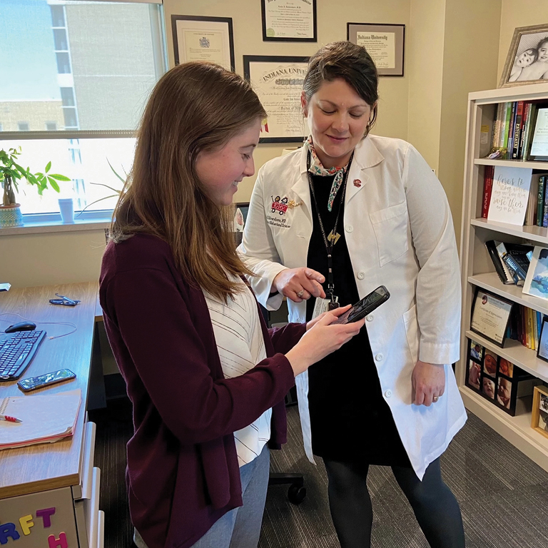 A woman wearing a doctor's lab coat smiles while pointing to a smartphone being held by a young woman standing to her left.