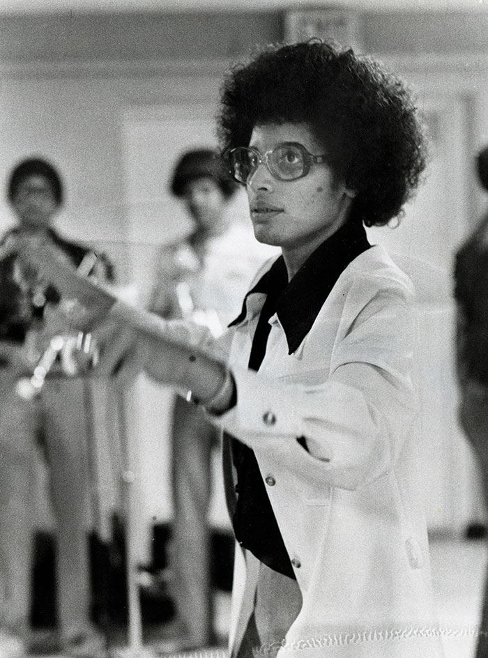 Black-and-white photo of a Black woman wearing a white leisure suit and glasses gestures with her hand while brass musicians stand in the background.