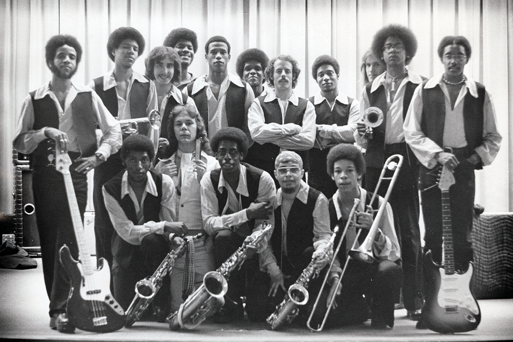 Black-and-white photo of an ensemble of sixteen musicians wearing matching shirts and vests and posing with their instruments.