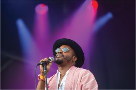 A Black singer wearing a black hat, sunglasses, and pink shirt holds the microphone while standing beneath purple spotlights.