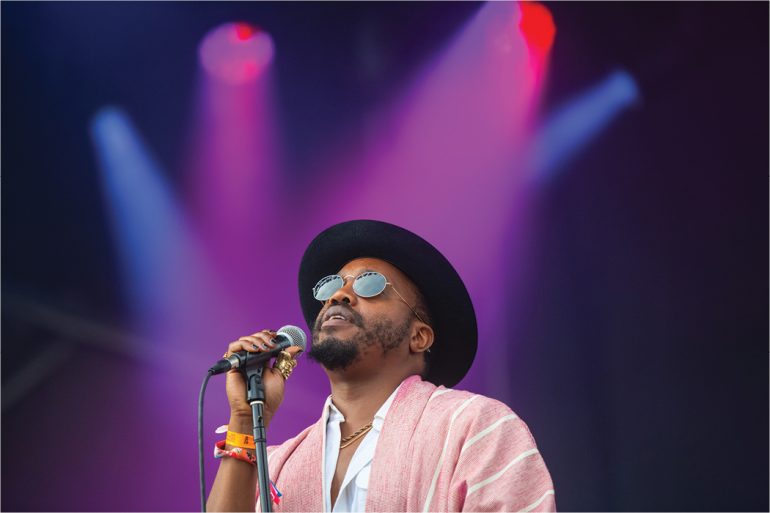 A Black singer wearing a black hat, sunglasses, and pink shirt holds the microphone while standing beneath purple spotlights.