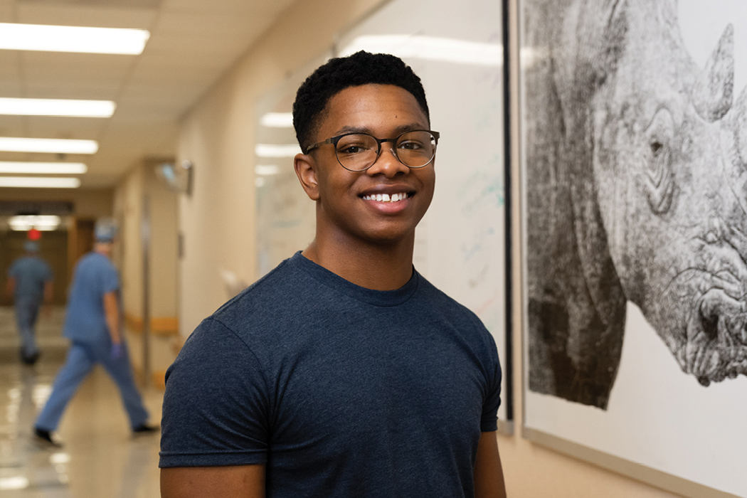 A young Black man wearing glasses and a blue T-shirt poses beside a dry-erase drawing of a rhinoceros in a hospital hallway.