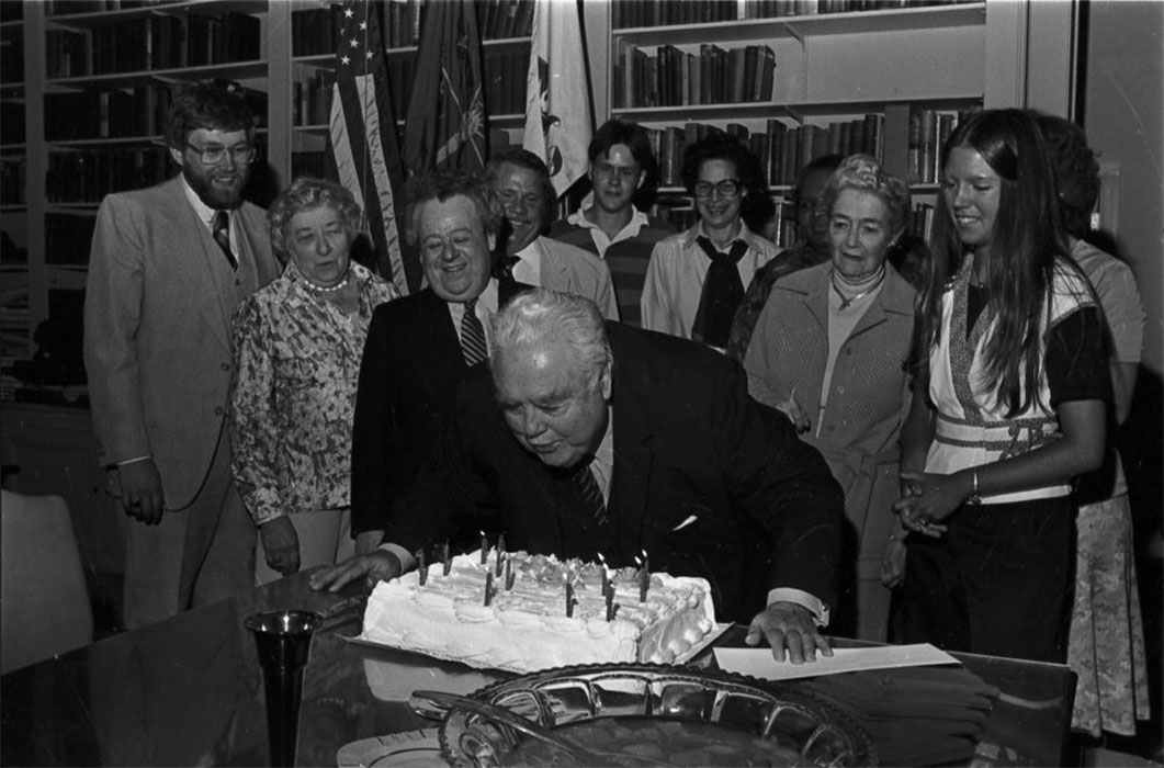 IU president Herman B. Wells blows birthday candles atop a cake surrounded by family and friends.