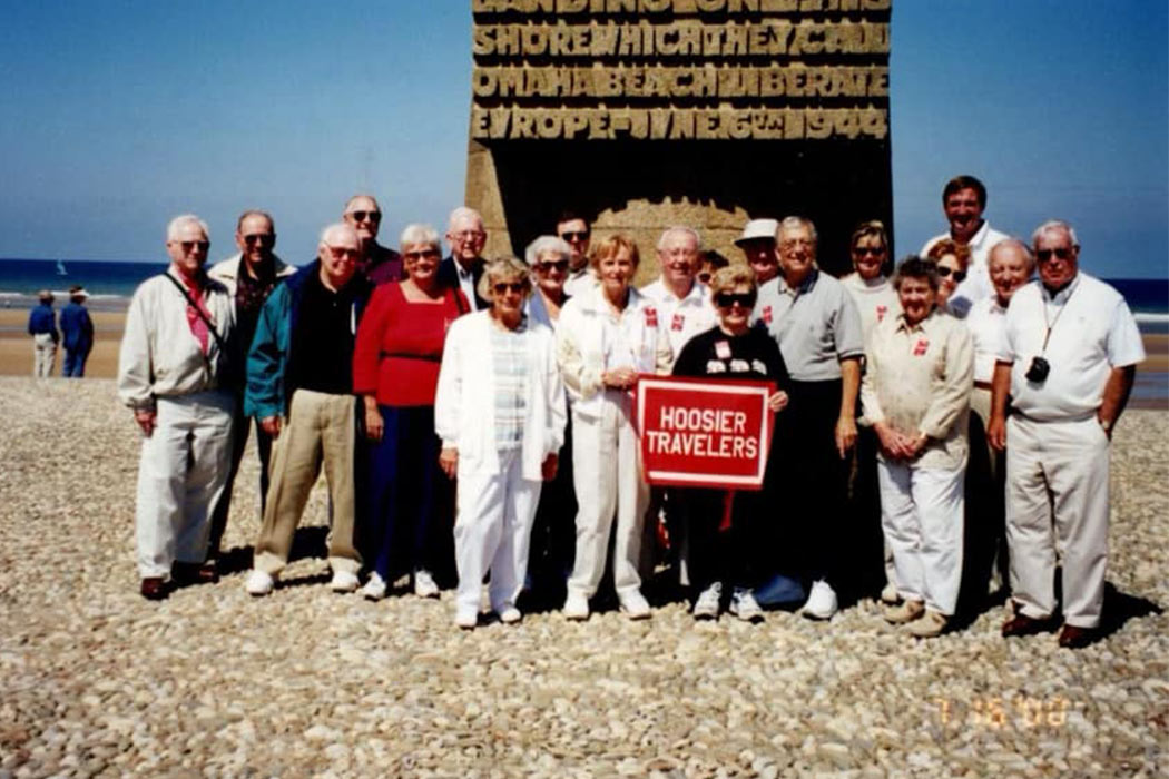 A group of people posing for a photo, in front stands Eleanor Cox Riggs holding a “Hoosier Travelers” sign.