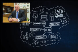 Tim Scales, wearing a suit and blue tie while seated behind his desk, overlaps a chalkboard illustration of a business plan that features the words "marketing," "strategy," "plan," "product," "success," and "idea."